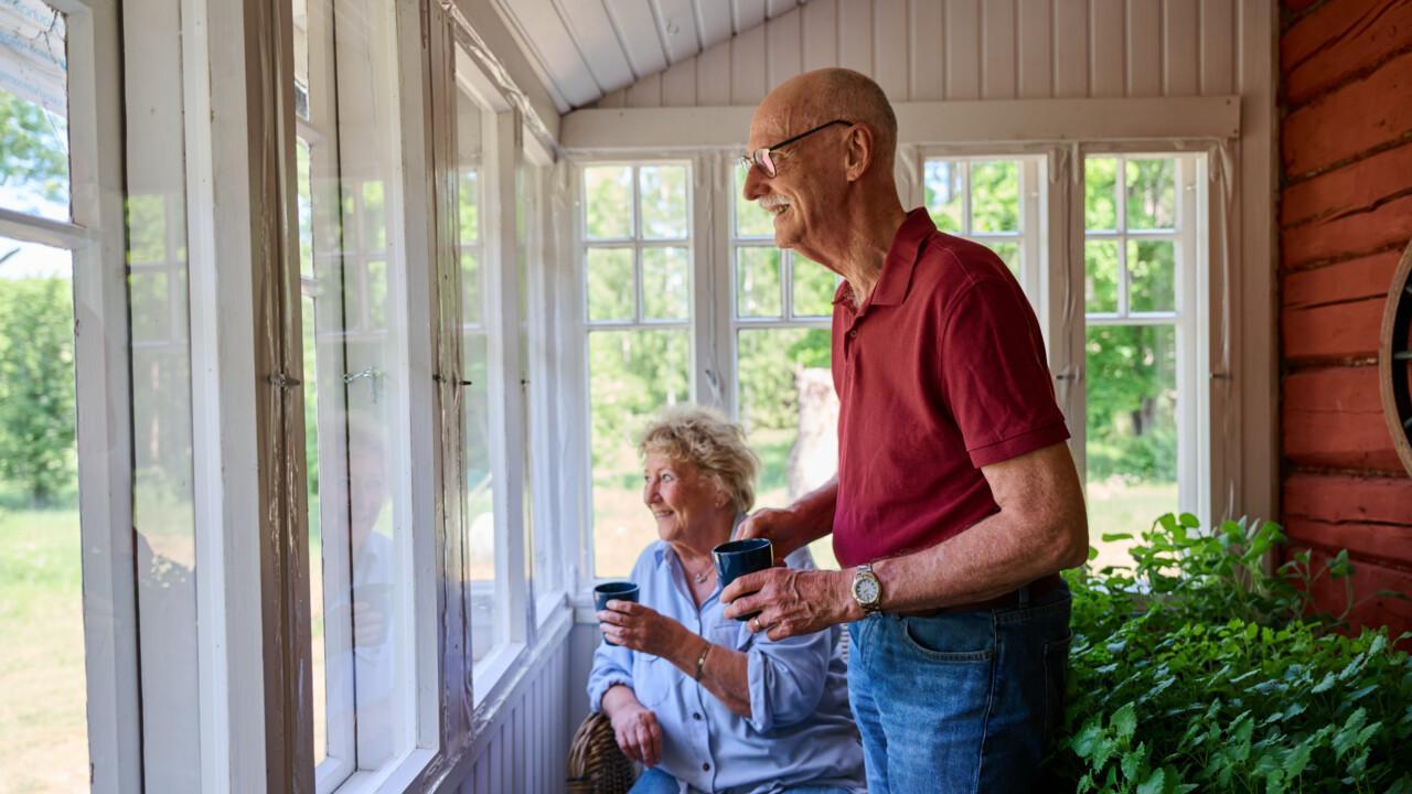 Iäkäs nainen istualtaan ja mies seisoen katselevat ikkunasta