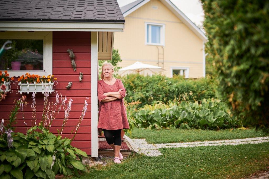 Iäkäs nainen seisoo punaisen piharakennuksen nurkalla käsivarret ristissä, taustalla vehreä puutarha ja keltainen omakotitalo.