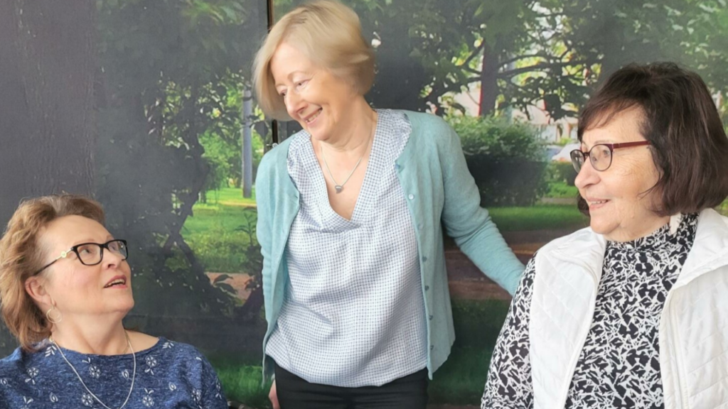 Kolme naista juttelee ja nauraa yhdessä sisätiloissa, taustalla on luontomaisemaa esittävä kuva.
