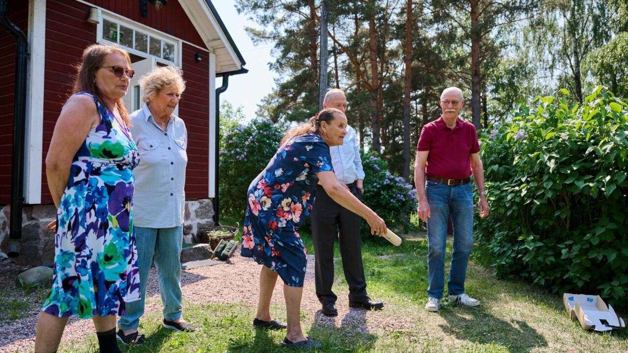kaksi iäkästä miestä ja kaksi iäkästä naista seisovat punaisen mökin edessä ja katsovat iäkkään naisen heittoa Mölkky-pelissä.