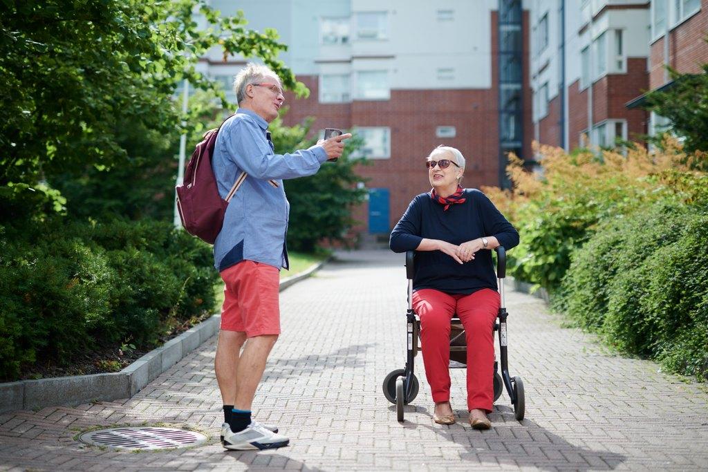 Mies ja nainen juttelevat ulkona, nainen istuu rollaattorin päällä, kerrostalon pihalla