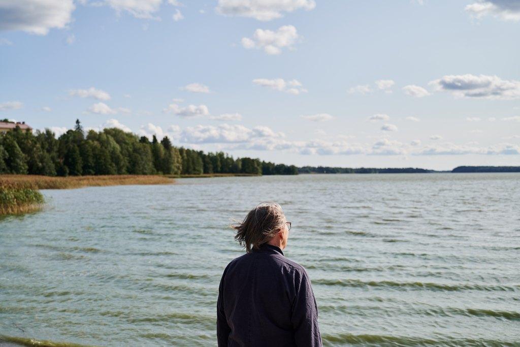 Harmaahiuksinen henkilö katselee edessään avautuvaa aaltoilevaa järvenselkää.