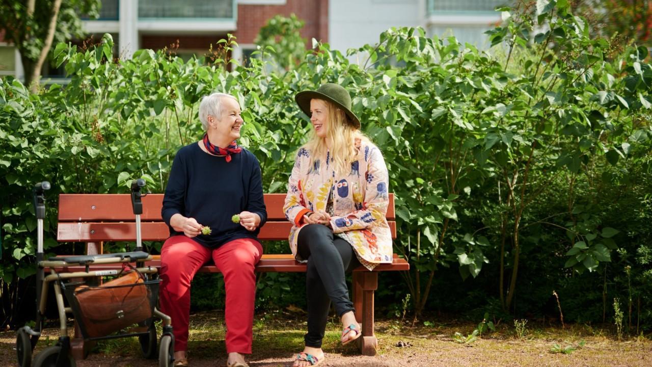 Vanha ja nuori nainen istuvat juttelemassa penkillä kerrostalon pihalla.