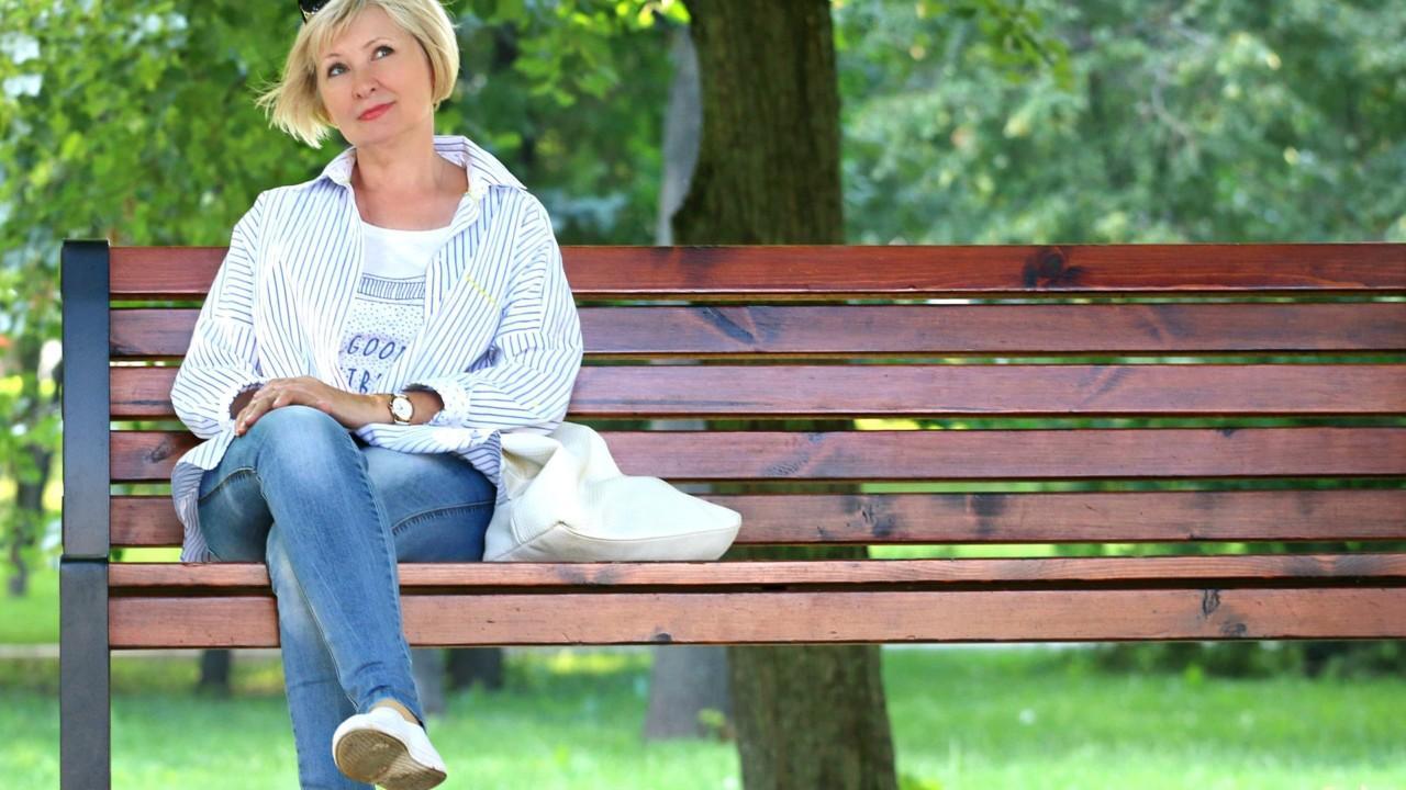 Keski-ikäinen nainen istuu yksin puistonpenkillä mietteliäänä.