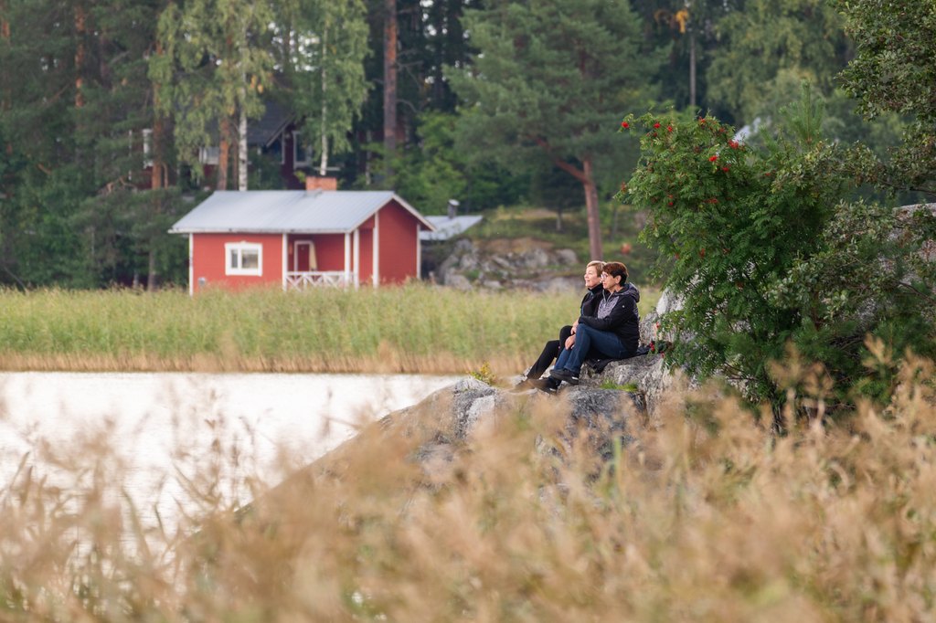 Mökki rannalla, ihmisiä istumassa kalliolla