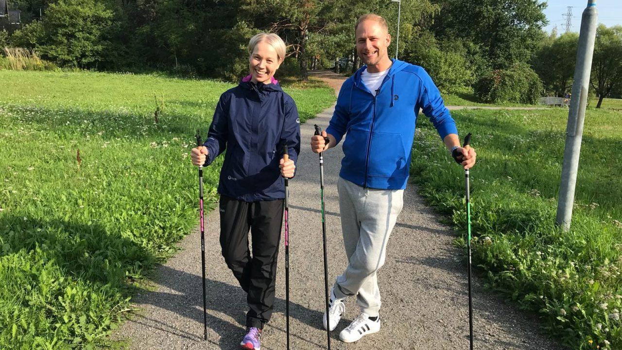 Nuori nainen ja mies seisovat ulkoiluvaatteissa kävelysauvat kädessä hiekkatiellä. Vihreää nurmikkoa ja metsää taustalla.