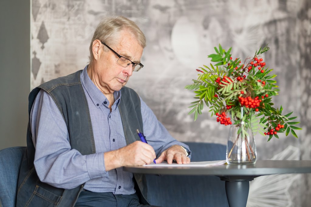 Silmälasipäinen iäkäs mies kirjoittaa kynällä paperille pöydän ääressä. Pöydällä on maljakossa pihlajanoksia ja punaisia marjoja.