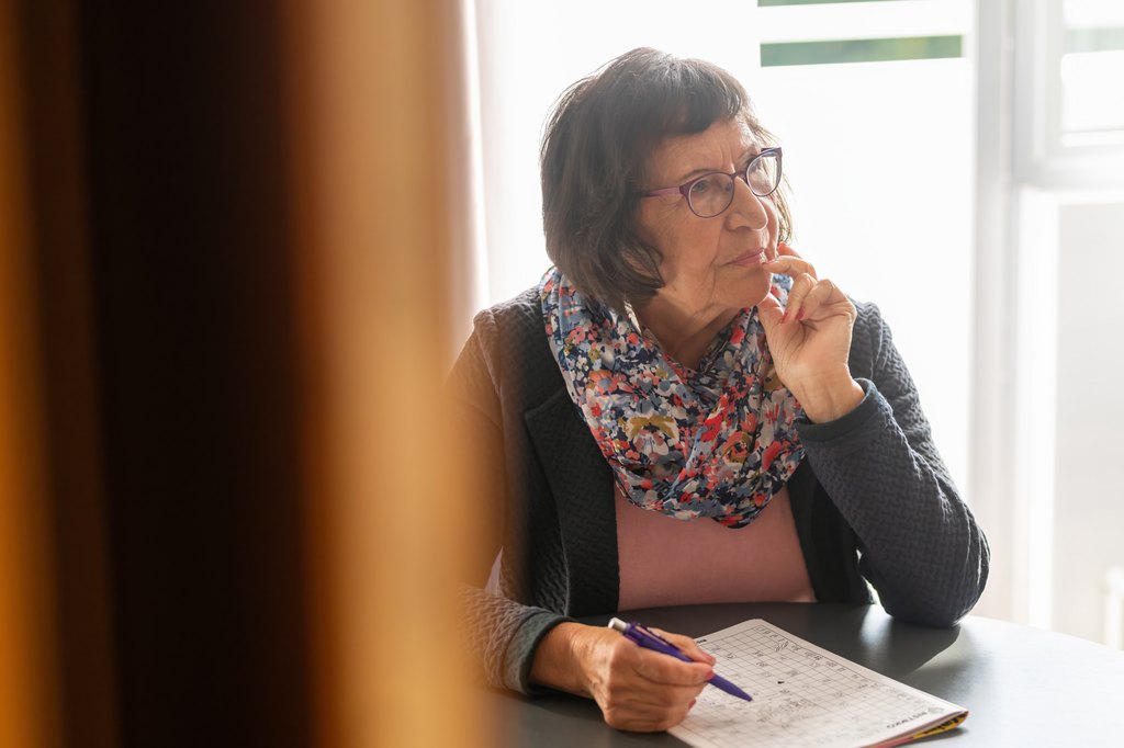 Silmälasipäinen iäkäs nainen istuu pöydän ääressä ja ratkoo kynä kädessään sanaristikkoa. Hän katsoo mietteliäänä sivulle.