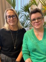Kaksi naista poseeraa kameralle sisätiloissa. Vasemmalla on vaaleahiuksinen nainen mustassa puserossa ja silmälaseissa, oikealla lyhyttukkainen nainen vihreässä neuleessa ja vihreäsankaisissa silmälaseissa. Taustalla on viherkasveja ja ikkuna.