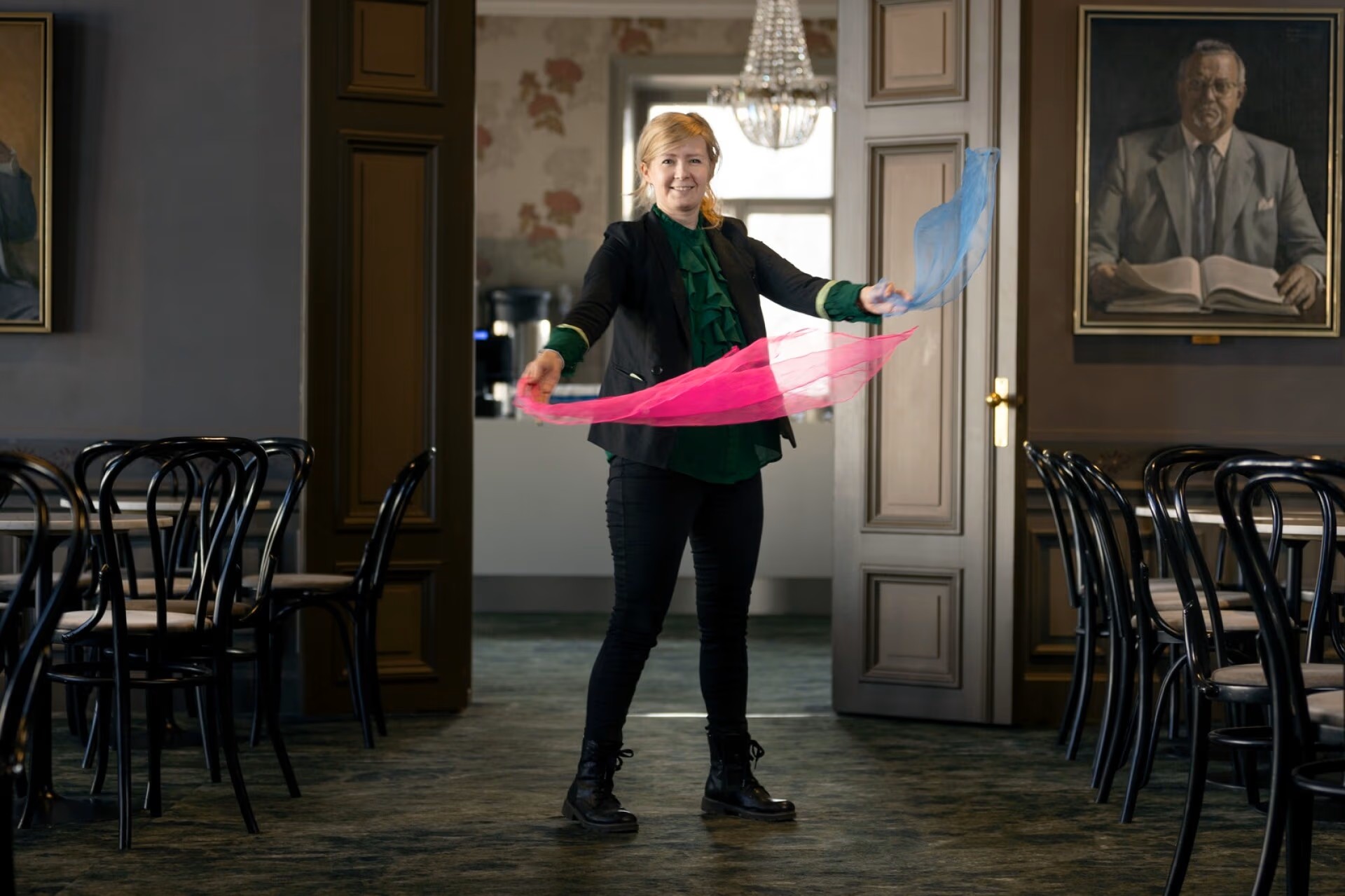Nainen seisoo juhlavassa salissa ja liikuttaa käsissään vaaleanpunaisia ja sinisiä huiveja. Ympärillä on pöytiä ja tuoleja, ja seinällä näkyy suuri muotokuva.